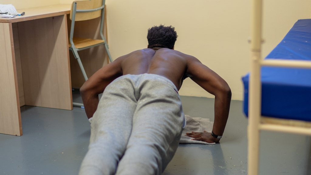 BME person in prison doing press ups on the floor with his top off in his cell.